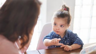 au centre de l'image une petite fille est assise à une table et à gauche de l'image une femme vue de dos est assise avec l'enfant
