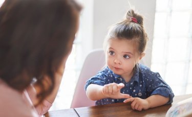 au centre de l'image une petite fille est assise à une table et à gauche de l'image une femme vue de dos est assise avec l'enfant