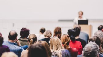au premier plan, des hommes et des femmes sont assis dans une salle de conférence. Ils écoutent une femme debout derrière un pupitre