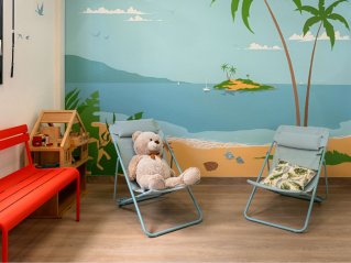 dans une pièce un ours en peluche est posé sur un petit transat devant un mur décoré  d'une image de plage