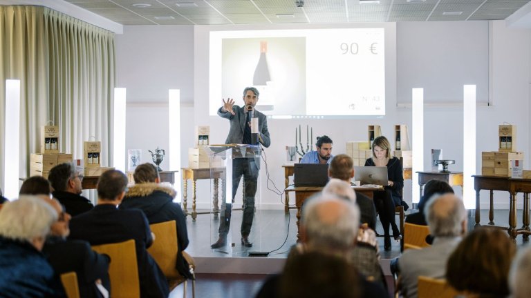 Dans une grande salle, un commissaire priseur anime une vente aux enchères devant un publict 