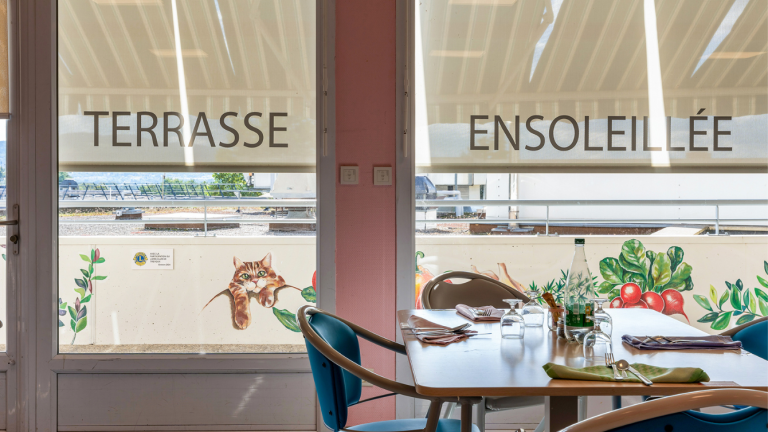 au premier plan une table avec quatre chaises. la table est mise. Au second plan une baie vitrée sur laquelle il est écrit terrasse ensoleillée
