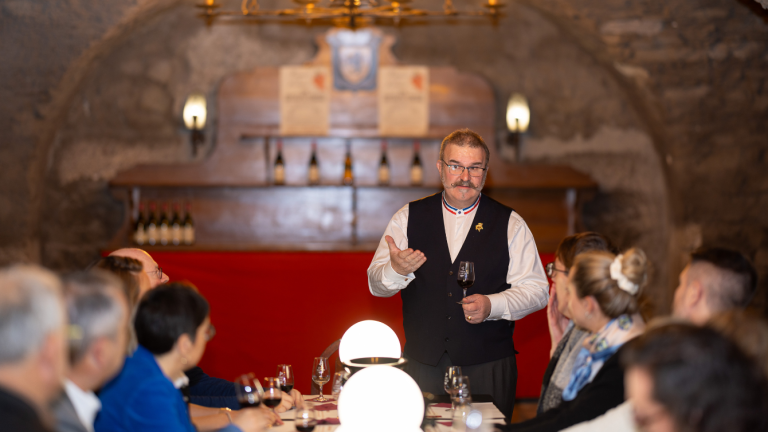 dans un caveau, un homme en chemise blanche et gilet noir parle debout à des personnes atablées