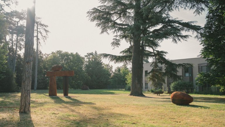 dans un parc, au premier plan une sculpture monumentale bois en forme de porte et au second plan un grand cèdre