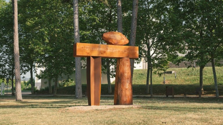 Dans un parc, au premier plan une grand sculpture bois en forme de porte, au second plan une allée de platanes