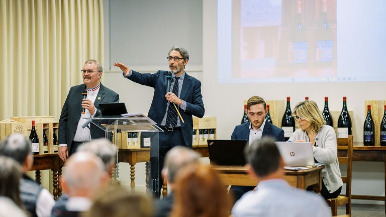 dans une grande salle, devant un pupitre un homme vêtu d'un costume lève le bras 