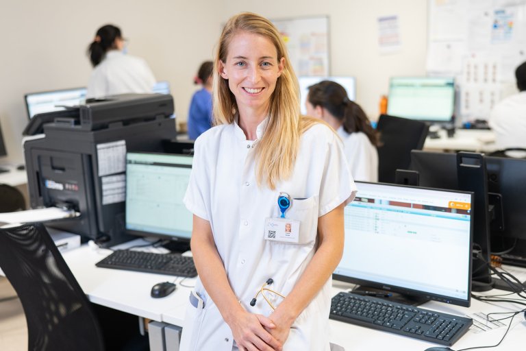 une jeune femme blonde, vêtue d'une blouse blanche est semi assise sur un bureau. Derrière elle, des écrans et des personnes qui sont devant leur poste de travail 