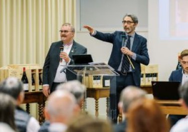 deux hommes en costume debout font face à un public assis, l'un des hommes lève le bras en direction du public